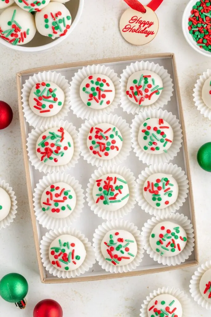 A box of white chocolate-covered cookie truffles, each in a cupcake liner, topped with red and green sprinkles. 