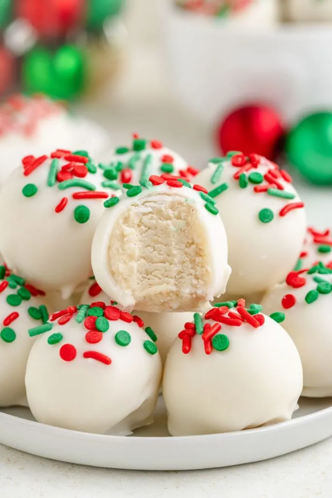 A plate of white truffles topped with red and green sprinkles. One truffle is partially eaten, revealing a creamy interior. Christmas ornaments are blurred in the background.