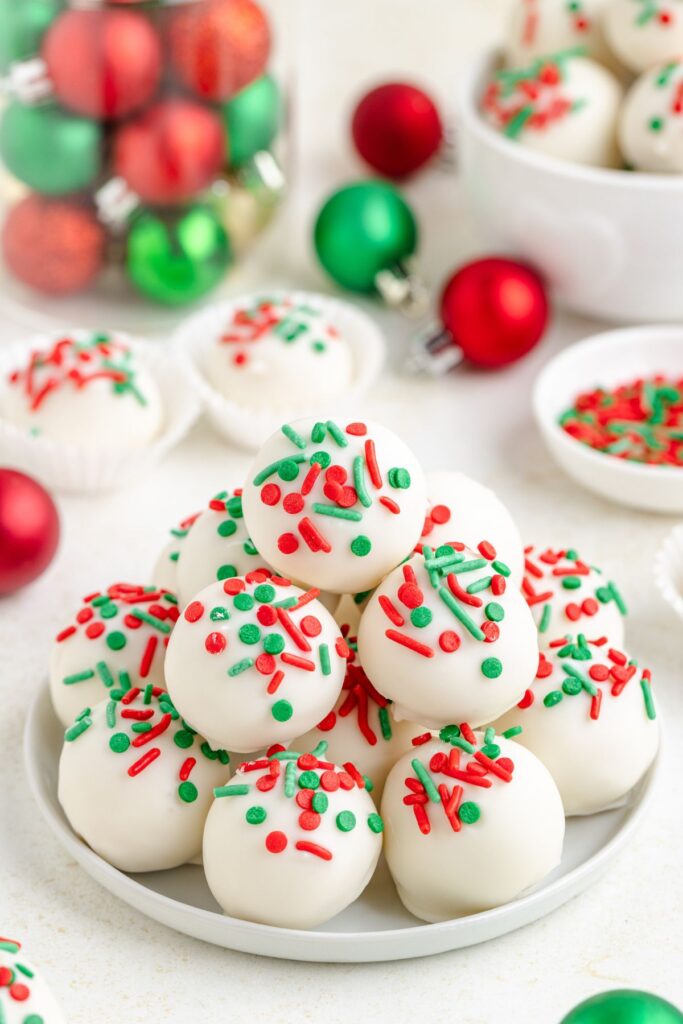 A plate of sugar cookie truffles decorated with red and green sprinkles, surrounded by small festive ornaments and additional truffles in the background, creating a holiday-themed setting.