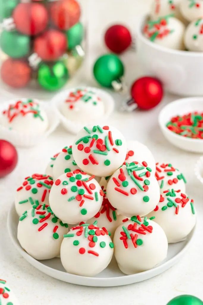 A plate of sugar cookie truffles decorated with red and green sprinkles, surrounded by small festive ornaments and additional truffles in the background, creating a holiday-themed setting.