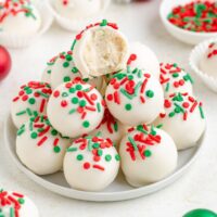 A plate of sugar cookie truffles, each topped with festive red and green sprinkles. One truffle is bitten, revealing a creamy interior. White paper cups and a bowl of sprinkles are in the background.