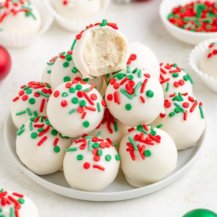 A plate of sugar cookie truffles, each topped with festive red and green sprinkles. One truffle is bitten, revealing a creamy interior. White paper cups and a bowl of sprinkles are in the background.