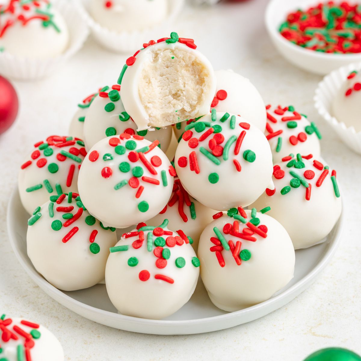 A plate of sugar cookie truffles, each topped with festive red and green sprinkles. One truffle is bitten, revealing a creamy interior. White paper cups and a bowl of sprinkles are in the background.