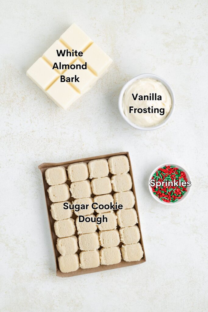 Top view of baking ingredients on a light background: a block of white almond bark, a container of vanilla frosting, a small bowl of red and green sprinkles, and a tray of sugar cookie dough squares.