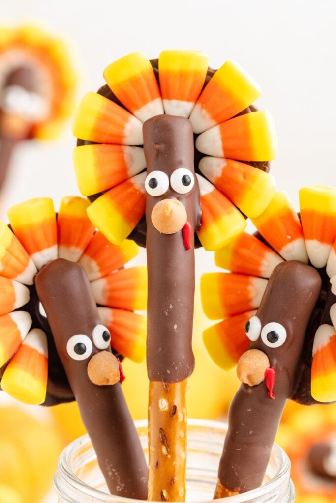 Three turkey-themed treats made of chocolate-covered pretzel rods with candy corn tail feathers and faces made of candy eyes, a chocolate nose, and a red candy wattle. They are displayed in a clear jar.