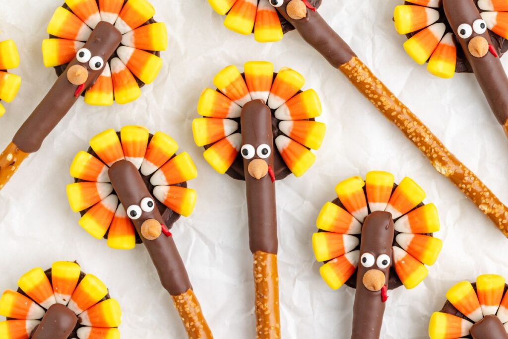 Pretzel sticks decorated as turkeys with candy corn and chocolate, laid out on a white surface. Each pretzel has candy corn feathers, a chocolate body, and candy googly eyes, creating a festive appearance.