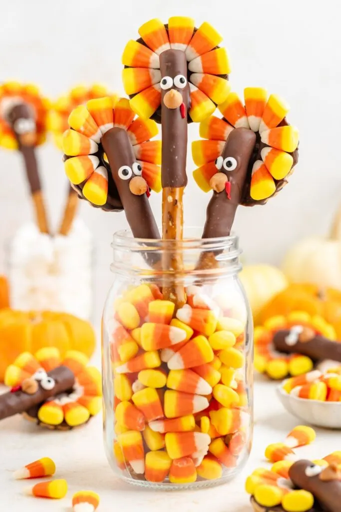 Candy corn turkeys on pretzel rods are arranged in a jar filled with candy corn. Each turkey has a chocolate-covered body, candy eyes, and a candy corn tail, with more turkeys and pumpkins blurred in the background.