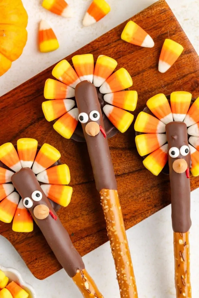 Three turkey-themed treats made from pretzel rods dipped in chocolate with candy corn for feathers and candy eyes, lying on a wooden board. Additional candy corn pieces are scattered around.