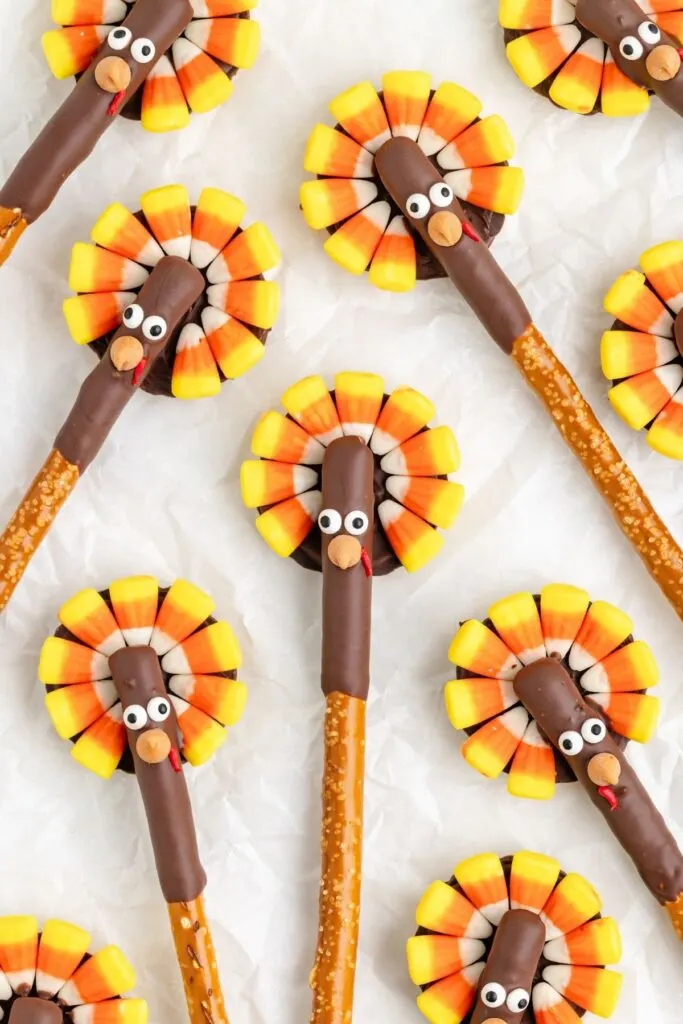 Pretzel sticks decorated with chocolate and candy corn to resemble turkeys, arranged on a white background. Each turkey has candy eyes and red icing for a beak. The candy corn forms tail feathers around the chocolate-covered pretzel tops.