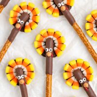 Pretzel sticks decorated with candy corn to resemble turkeys are arranged on white paper. Each stick is partially covered in chocolate, with candy eyes and a small orange candy for the beak, surrounded by candy corn resembling tail feathers.