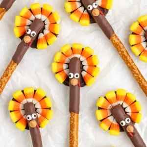 Pretzel sticks decorated with candy corn to resemble turkeys are arranged on white paper. Each stick is partially covered in chocolate, with candy eyes and a small orange candy for the beak, surrounded by candy corn resembling tail feathers.