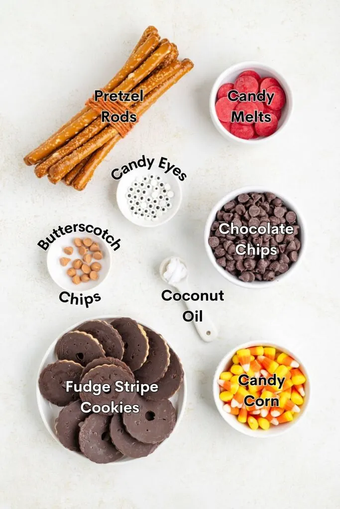 A flat lay of baking ingredients, including pretzel rods, candy melts, candy eyes, butterscotch chips, chocolate chips, coconut oil, fudge stripe cookies, and candy corn, each labeled in bowls or plates.