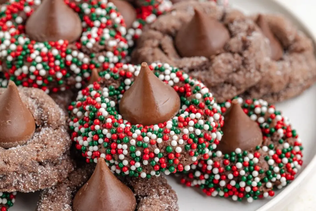 Close-up of chocolate cookies topped with chocolate candy kisses, decorated with red, green, and white sprinkles, arranged on a white plate.