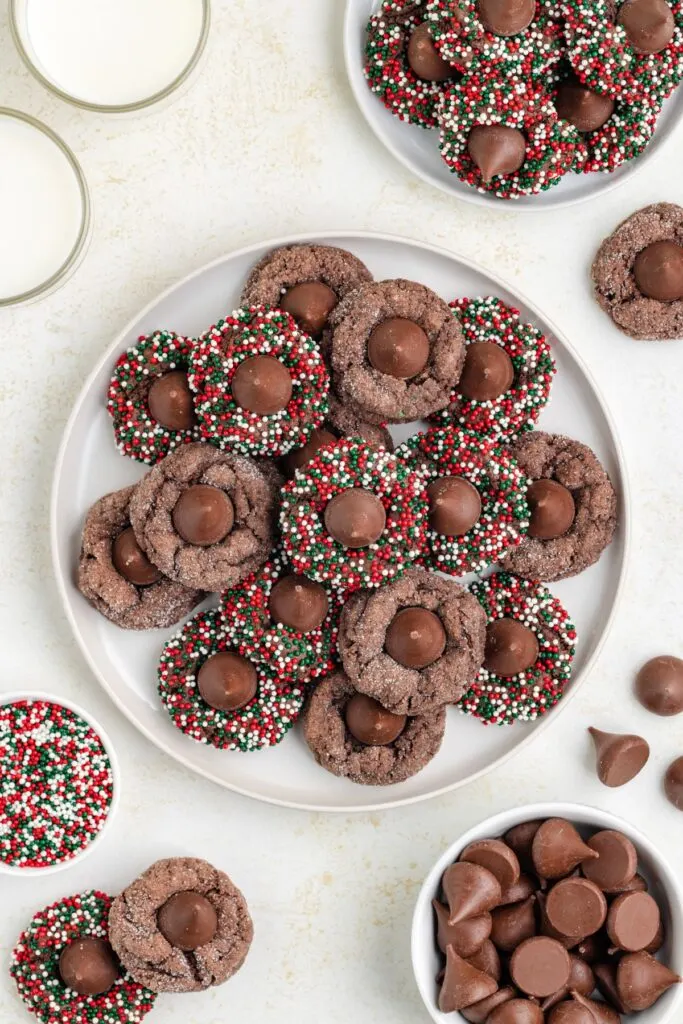 A plate piled with chocolate kiss cookies, surrounded by sprinkles in festive red, green, and white colors. Additional cookies, chocolate drops, sprinkles, and glasses of milk are set around the plate.