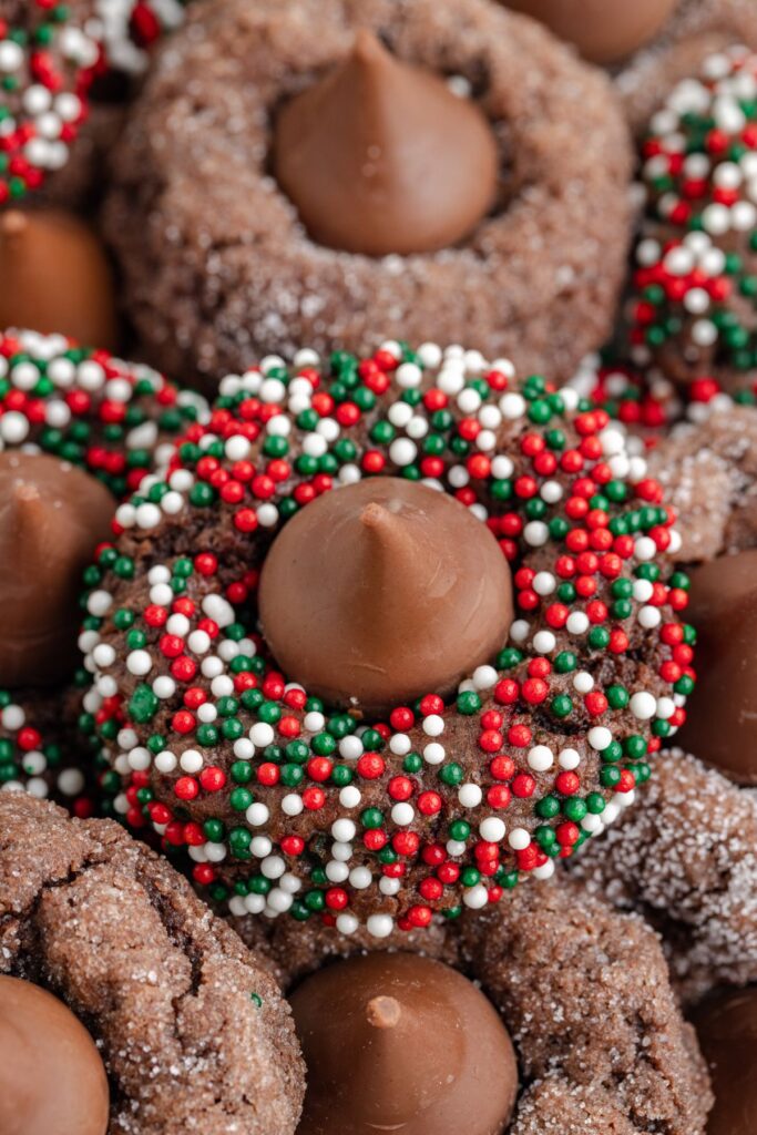 Chocolate kiss cookies topped with Hersheys Kisses, some decorated with red, green, and white sprinkles, are stacked together. The cookies textures are rich and inviting.