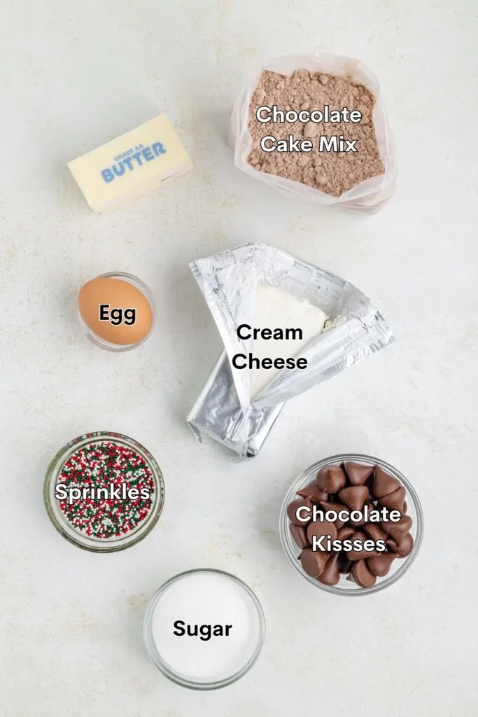 Ingredients for baking including a stick of butter, chocolate cake mix in a bag, an egg, cream cheese in foil, a jar of multicolored sprinkles, a bowl of sugar, and a bowl of chocolate kisses arranged on a light surface.