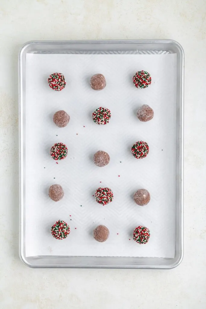 A baking tray with 15 chocolate dough balls arranged in a grid pattern on parchment paper. Some balls are coated in colorful sprinkles, while others are plain with a light dusting of sugar.