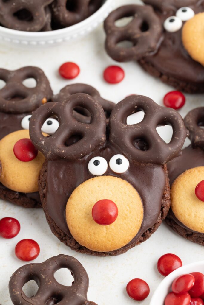 Chocolate reindeer cookies with round cookies for noses, red candies as accents, chocolate-covered pretzels for antlers, and white candy eyes. Red candies scattered around add a playful touch to this festive treat.