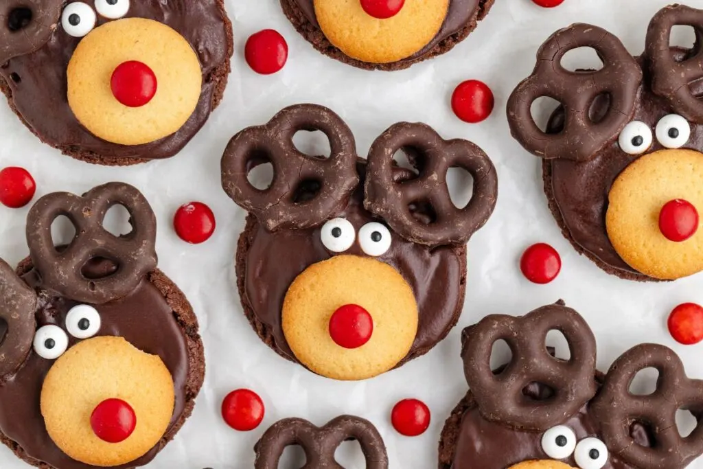 Festive reindeer-themed chocolate cookies with candy eyes, vanilla wafer snouts, red candy noses, and pretzel antlers are arranged on a white surface, surrounded by scattered red candies.