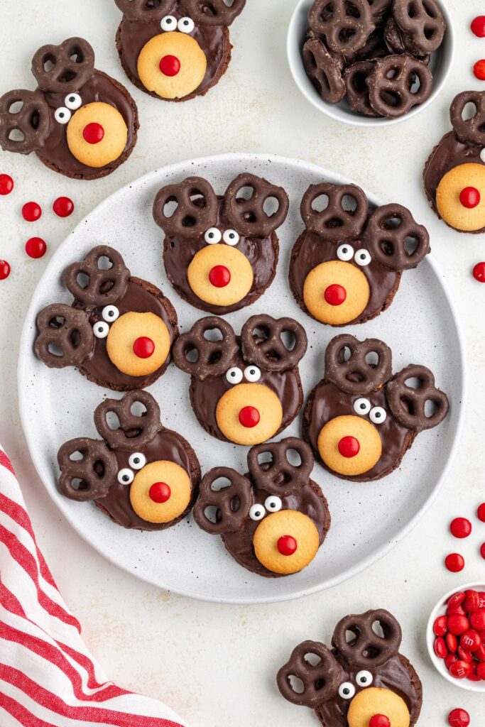 A plate of reindeer-themed chocolate cookies, with pretzels for antlers, red candies for noses, and white candy eyes. More cookies and candy are scattered around the plate. A red-striped cloth is visible at the edge.