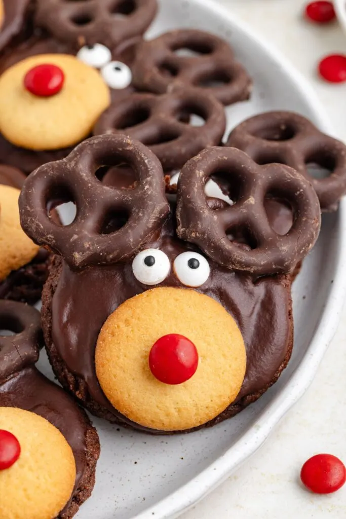 Chocolate cookies decorated to look like reindeer with chocolate-covered pretzels as antlers, candy eyes, a beige cookie for a snout, and a red candy nose. Placed on a white plate with red candies scattered around.