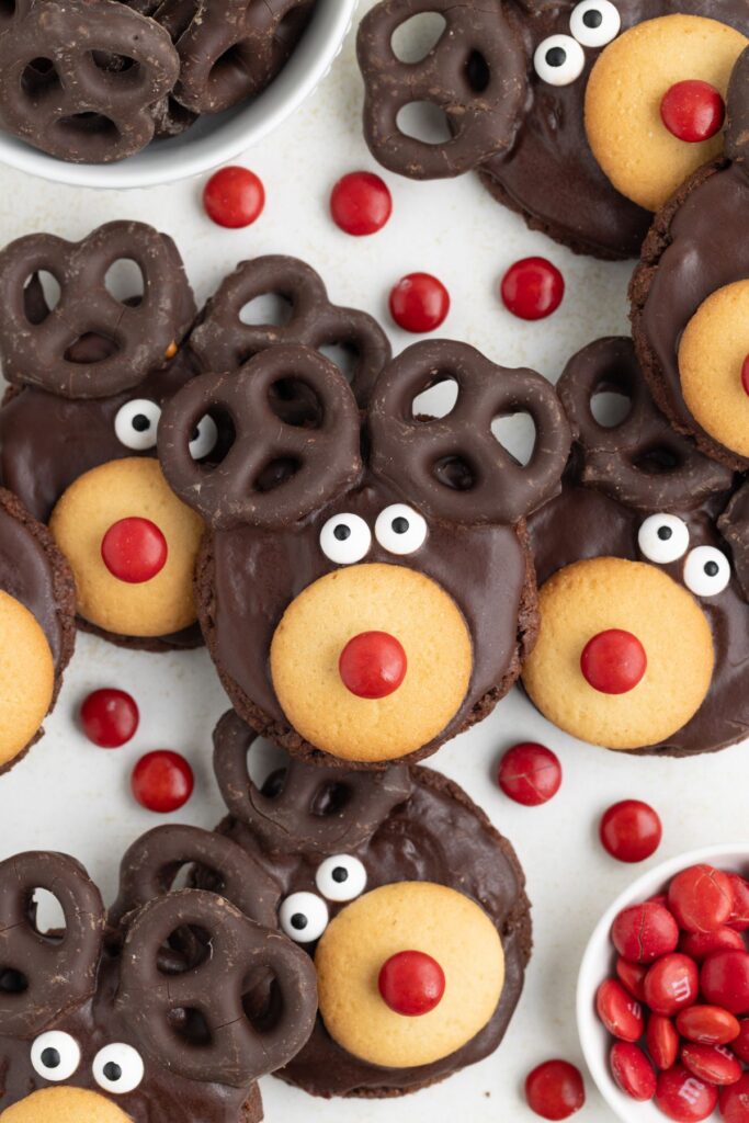 Cookies decorated as reindeer with chocolate-covered pretzels for antlers, vanilla wafer snouts, candy eyes, and red candy noses. Surrounded by red candies and extra pretzels.