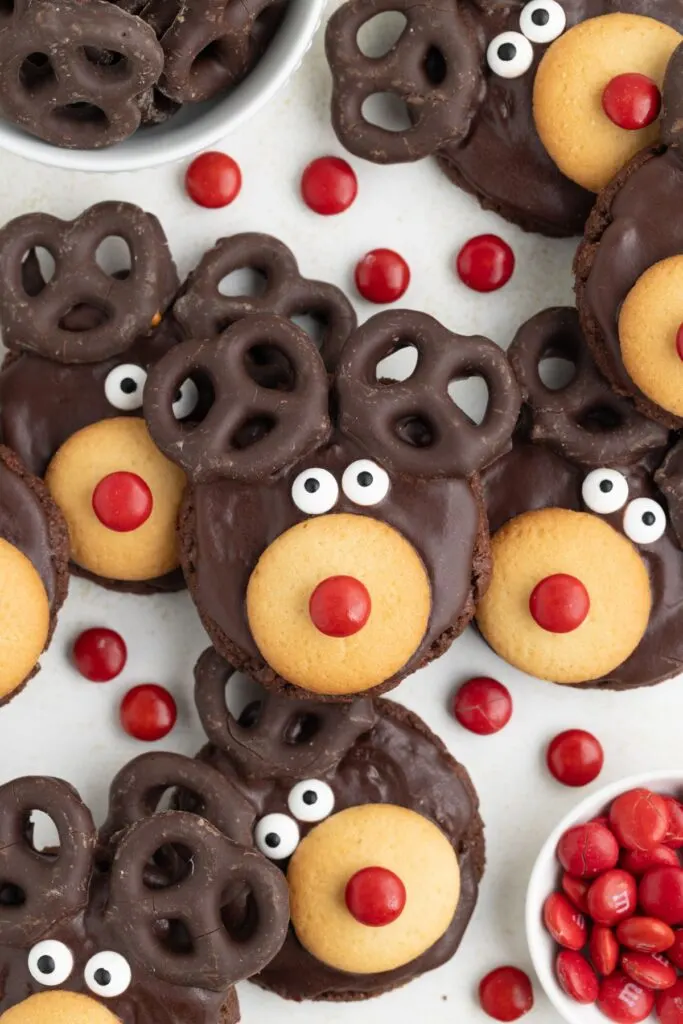 Cookies decorated as reindeer with chocolate-covered pretzels for antlers, vanilla wafer snouts, candy eyes, and red candy noses. Surrounded by red candies and extra pretzels.