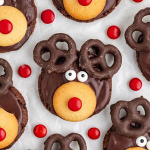 A festive cookie designed to resemble a reindeer, with chocolate-covered pretzels as antlers, candy eyes, a red candy nose, and a vanilla wafer for the snout. Surrounded by red candies on a white background.