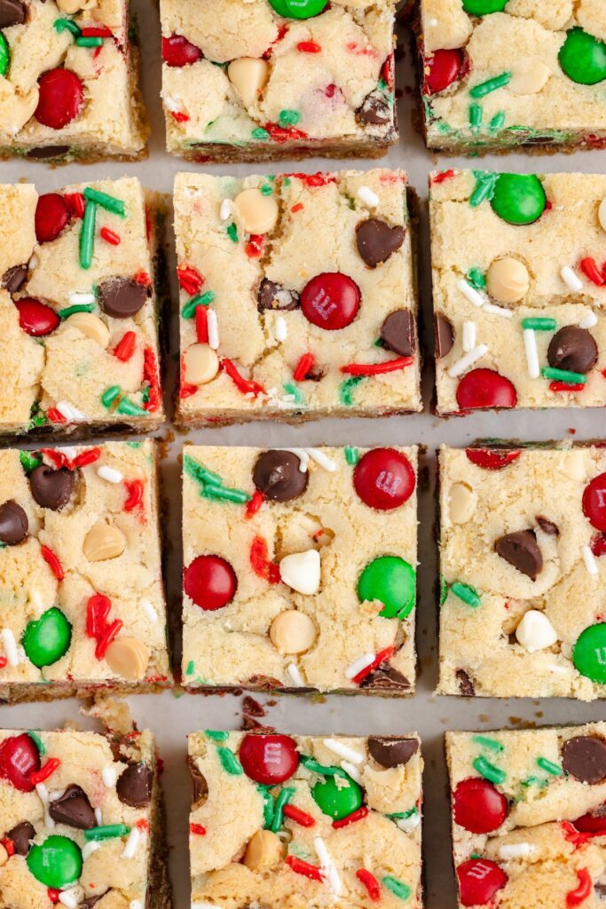 Sliced cookie bars topped with red and green candies, chocolate chips, and sprinkles are arranged in a grid pattern. The bars have a soft, golden-brown appearance with festive holiday decorations.