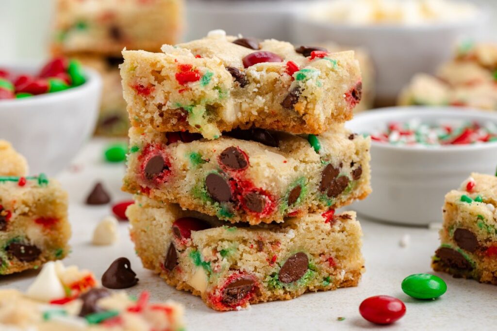 A stack of Christmas cookie bars filled with red and green candy-coated chocolates and chocolate chips. The bars are surrounded by more candy pieces in a bright, colorful setting.