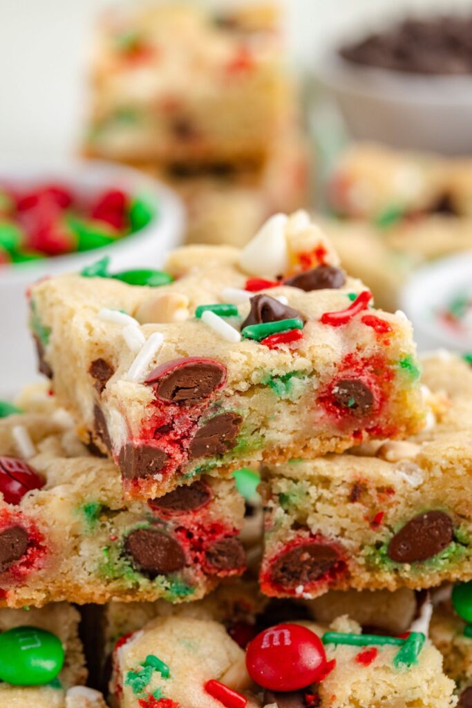 A stack of Christmas cookie bars topped with red, green, and white chocolate candies, along with dark chocolate chips. The background features bowls containing more candies and chocolate chips.