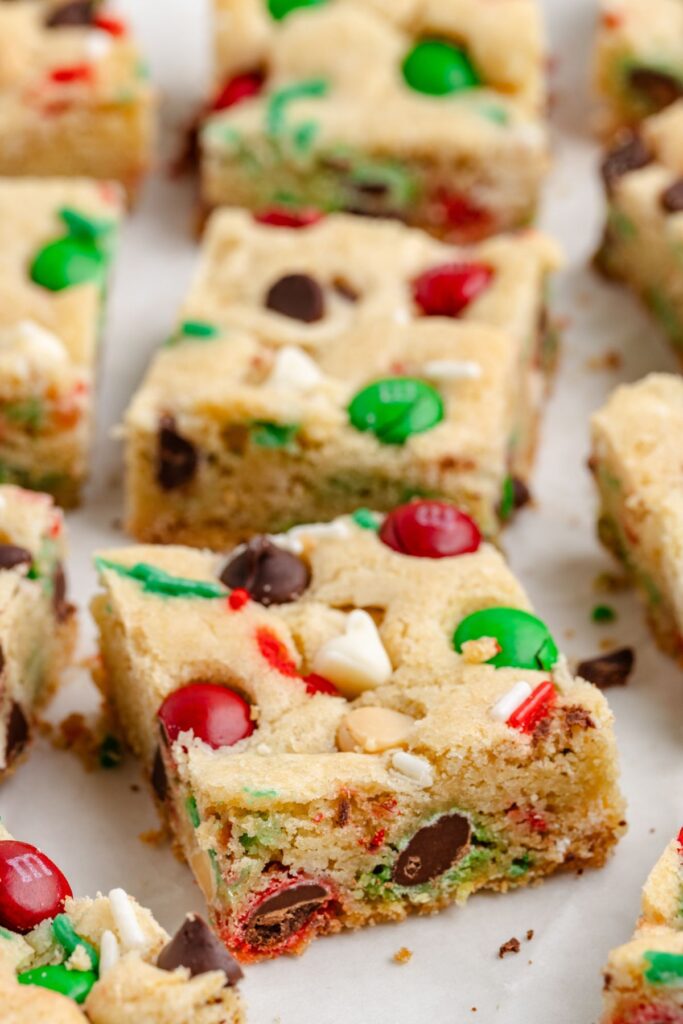 Close-up of holiday-themed cookie bars with red and green candies, chocolate chips, and white chocolate chunks. The bars are cut into squares and arranged on a white parchment paper.