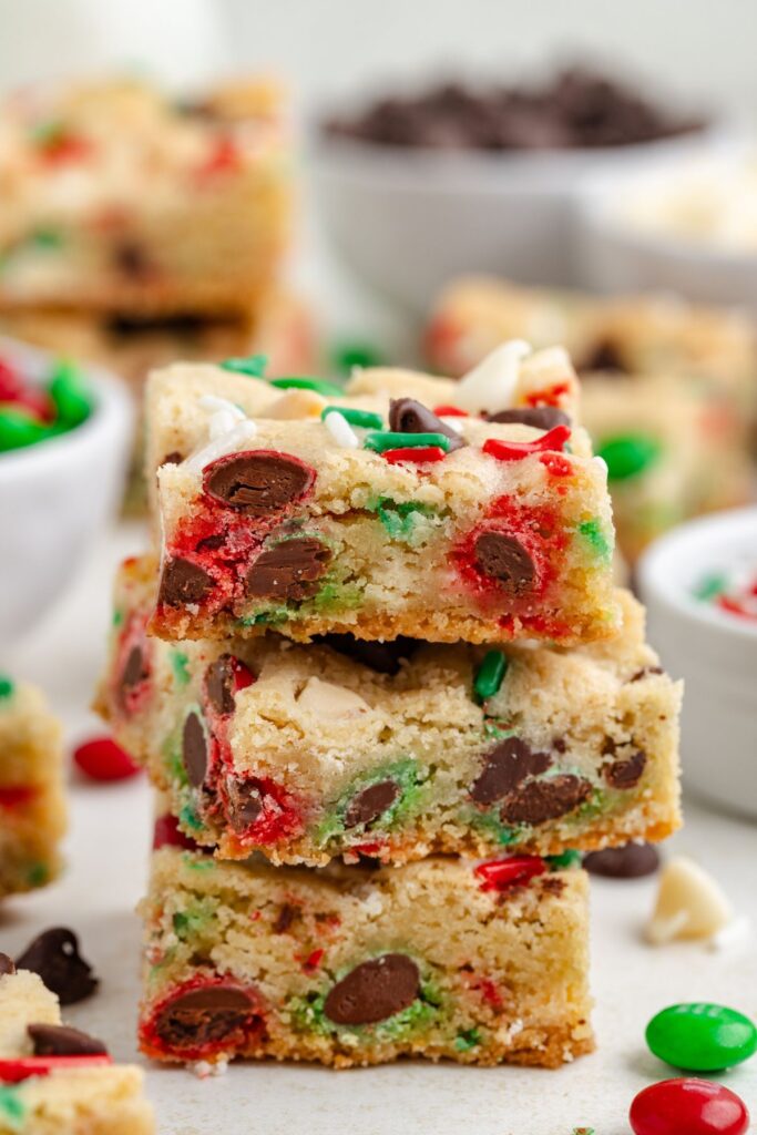 Three stacked Christmas cookie bars with colorful red, green, and chocolate candies are displayed. Blurred in the background are bowls filled with more candies and a few scattered around the table.