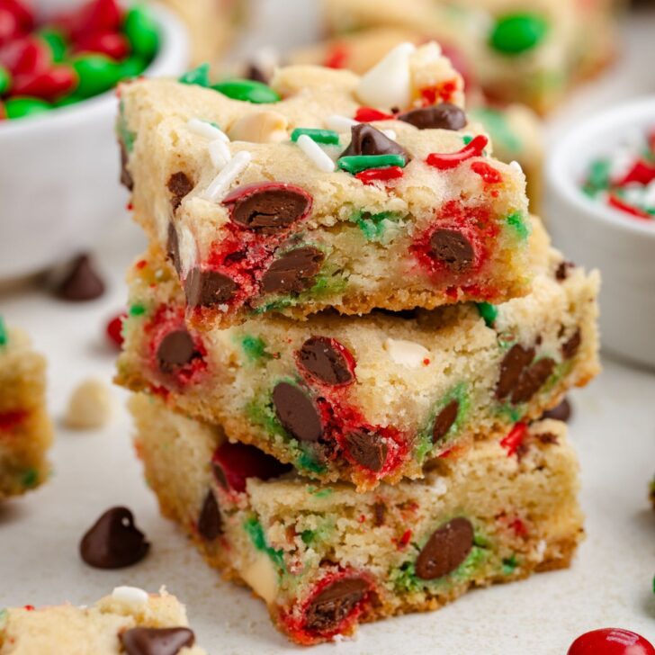 A stack of Christmas cookie bars with chocolate chips, red and green candies, and sprinkles sits on a white surface. Scattered candies and chocolate chips are visible in the background, adding to the festive scene.