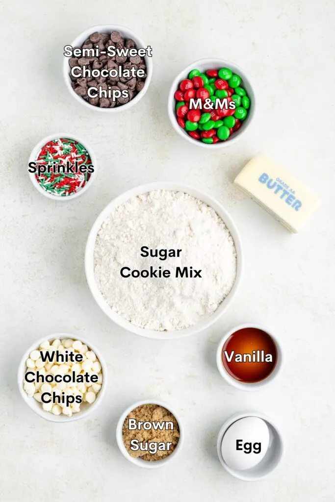 Image of baking ingredients including a bowl labeled Sugar Cookie Mix, surrounded by semi-sweet chocolate chips, M&Ms, sprinkles, white chocolate chips, vanilla, brown sugar, an egg, and a stick of butter.