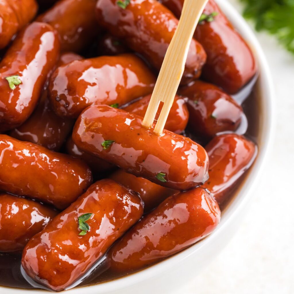 A close-up of cocktail sausages coated in a glossy grape jelly and barbecue sauce, garnished with small parsley bits. A wooden skewer is picking up one sausage from a white bowl.