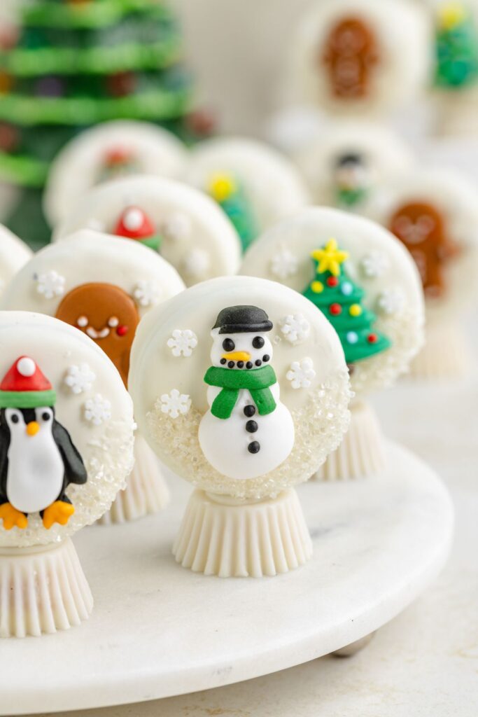 Oreo snow globes decorated with winter themes on a white platter. Designs feature a snowman, Christmas tree, and penguin, all nestled among snowflake sprinkles. 