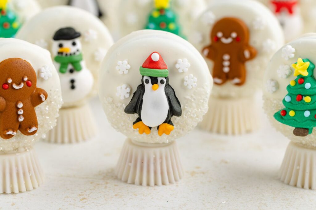 Holiday-themed Oreo snow globes with intricate decorations: A penguin wearing a red hat, surrounded by snowflakes, features prominently. Nearby, gingerbread figures, snowmen, and Christmas trees add to the festive display, all on white peanut butter cup bases.