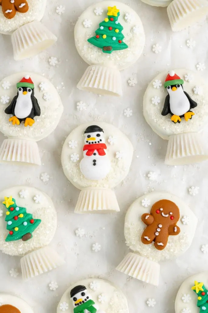 Festive Oreo snow globe cookies decorated with icing: penguins in red hats, Christmas trees, snowmen, and gingerbread men. Theyre placed on a white surface with small snowflake decorations scattered around.