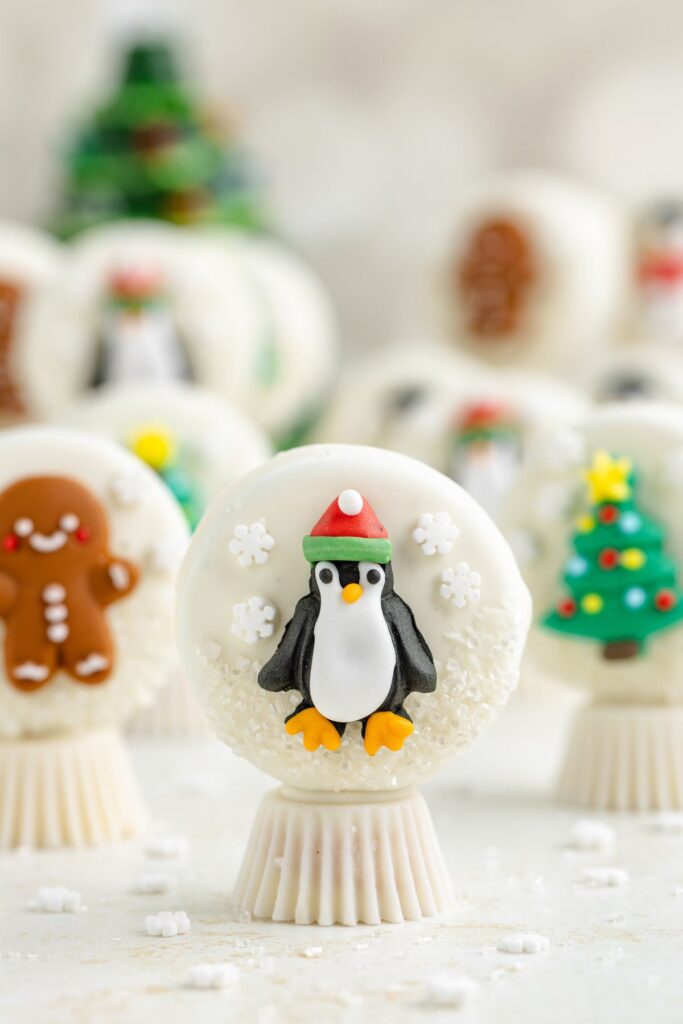 A festive white chocolate cake pop decorated with a penguin wearing a Santa hat, surrounded by snowflakes. Other cake pops with gingerbread man and Christmas tree designs are blurred in the background.