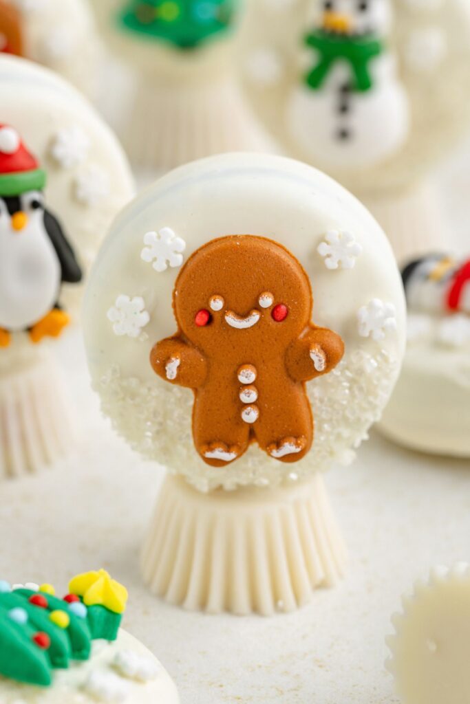 Oreo Snow Globes featuring a smiling gingerbread man on white chocolate Oreo, surrounded by edible snowflakes nestled among other holiday-themed treats in the background.