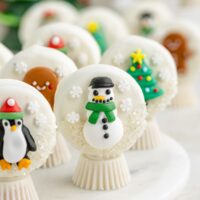 Oreo snow globes decorated with icing characters. The foreground features a white chocolate-covered cookie topped with a snowman design. Other cookies display a penguin, gingerbread figure, and Christmas tree, all with colorful icing details.