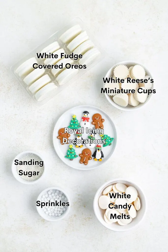 A top view of various holiday-themed baking ingredients: white fudge covered Oreos, white Reeses miniature cups, royal icing decorations, sanding sugar, sprinkles, and white candy melts arranged in bowls on a light surface.