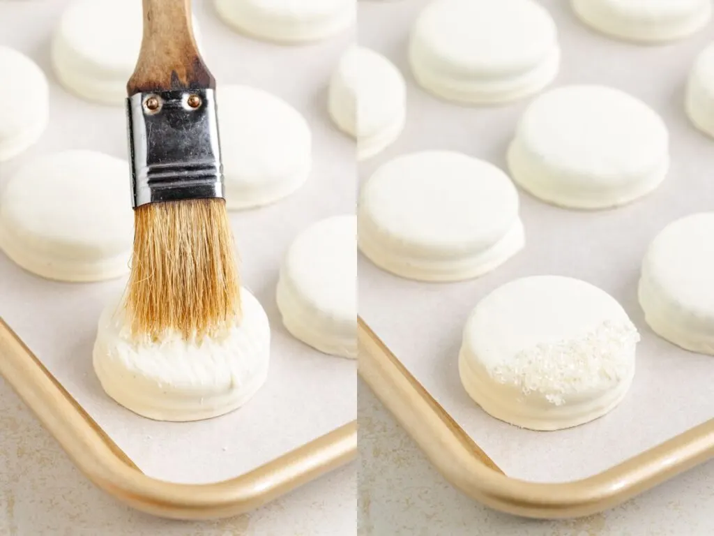 A close-up of a baking tray with white chocolate Oreos. On the left, a brush is applying melted white chocolate to an Oreo. On the right, the same Oreo is sprinkled with sugar, placed on parchment paper.