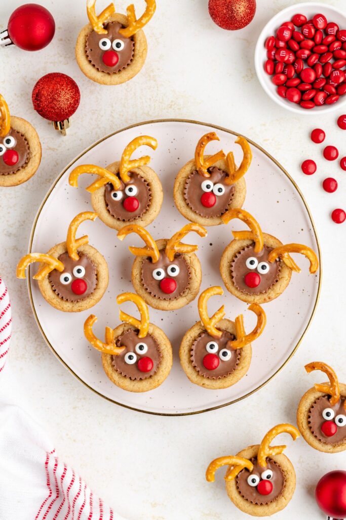 A plate of reindeer-themed cookies with red noses, white eyes, and pretzel antlers. Scattered red candies and a festive red ornament surround the plate on a white background.