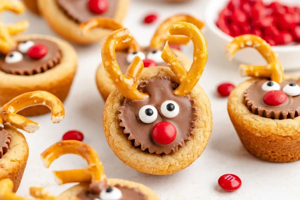 Close-up of reindeer-themed cookies with pretzel antlers, candy eyes, and red noses on a light surface. A bowl of red candies is in the background. Cookies are made of peanut butter cups and topped with chocolate.