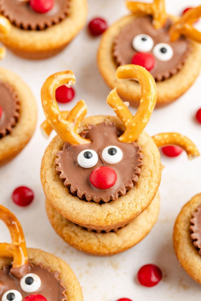 Cookies decorated to look like reindeer with pretzel antlers, chocolate peanut butter cups, candy eyes, and red candy noses are arranged on a white surface, surrounded by a few loose red candies.