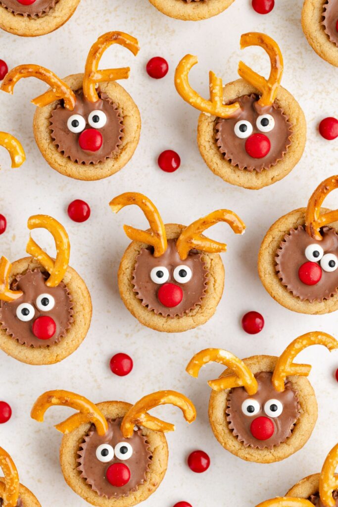 Reindeer-themed cookies decorated with red candy noses, pretzel antlers, and candy eyes are arranged on a light surface. Scattered around are red candies, enhancing the festive pattern.