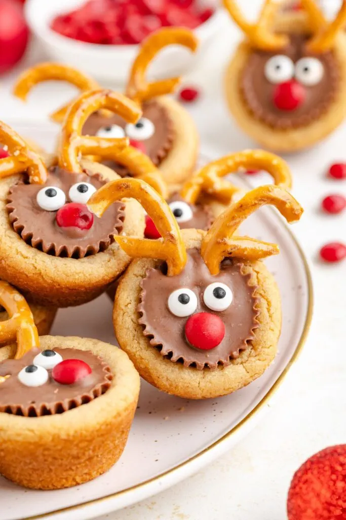 A plate of festive reindeer cookies, each topped with a chocolate candy, googly eyes, a red nose, and pretzel antlers. There are red candies and a white bowl in the blurred background.