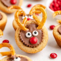 A Peanut Butter Cup Reindeer cookie cup with pretzel antlers, candy eyes, and a red candy nose is nestled among red candies and similar cookies on a white background.